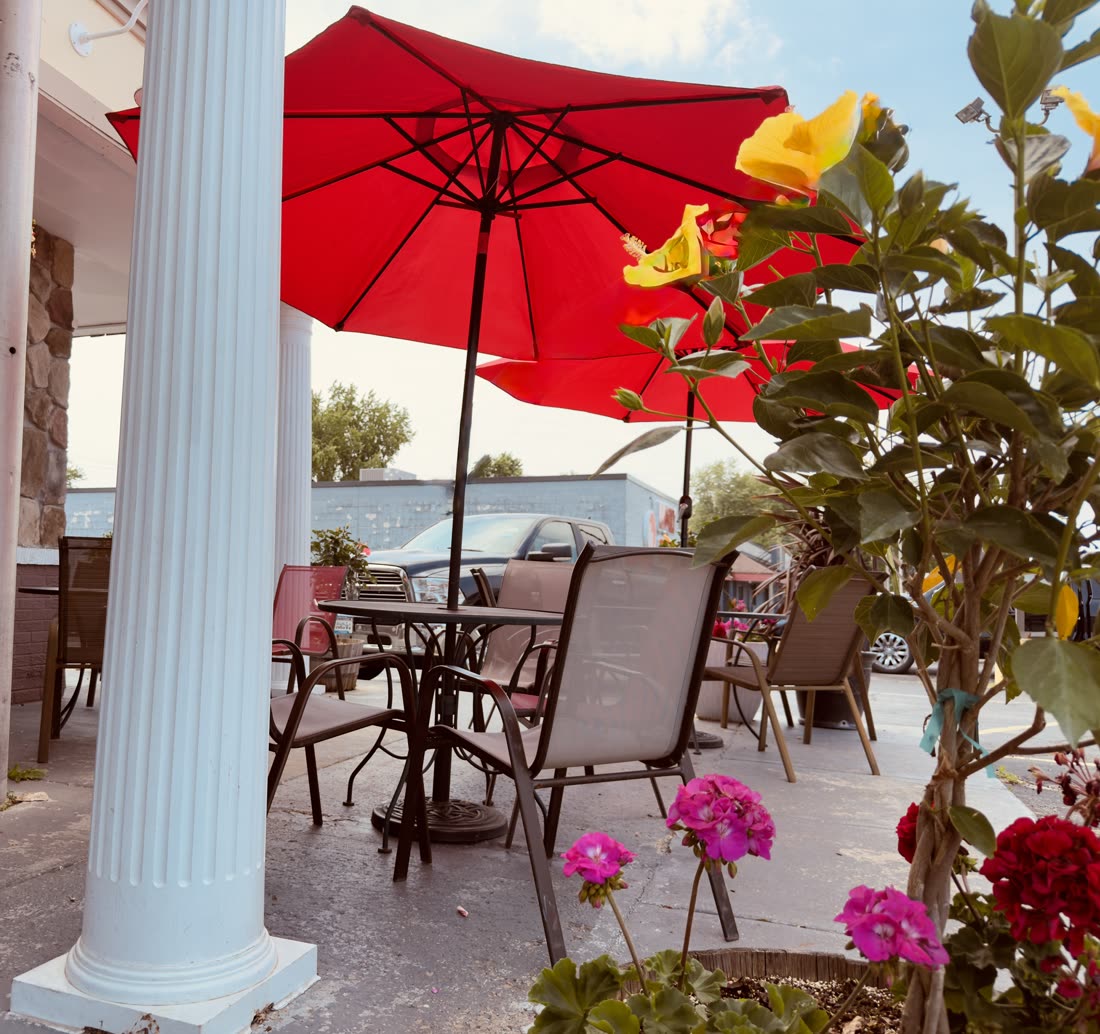 Outdoor patio seating between the white columns at Roman Castle, with red umbrellas and summer flowers